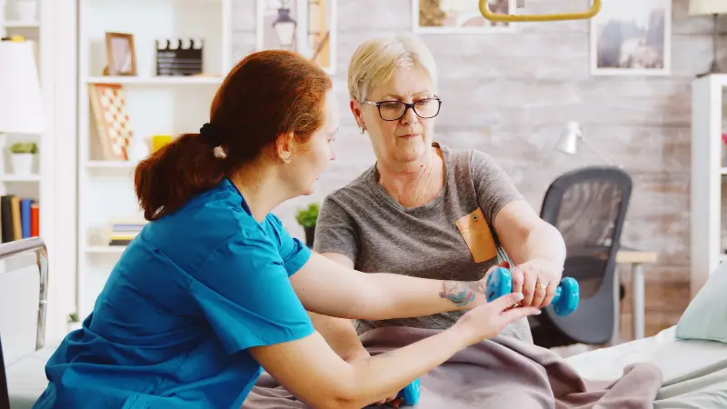 Eine einfühlsame Krankenschwester unterstützt eine ältere Frau bei der Muskelrehabilitation nach einem Unfall. Die Patientin liegt in einem Pflegebett in einem Seniorenheim, während die Krankenschwester ihr behutsam bei Bewegungsübungen hilft.